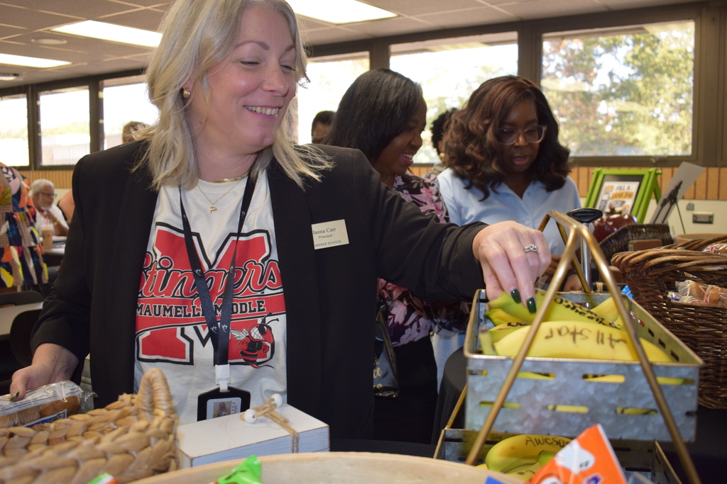 A principal wearing a black blazer and a Maumelle Middle shirt smiling as she reaches into a basket to grab a banana marked with an encouraging message. Other colleagues stand in the background near a table filled with breakfast items and snacks.
