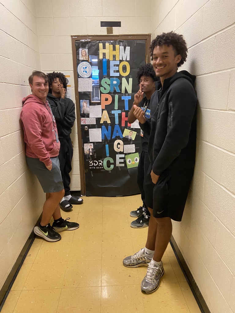 Four high school students posing in a hallway beside a classroom door decorated with colorful lettering and profiles of Hispanic entrepreneurs for Hispanic Heritage Month.