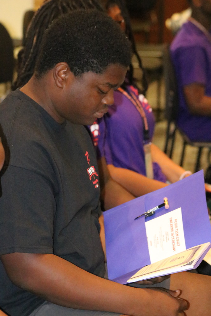 A student reads a paper within a folder. Inside is a program for Teen Court, a gavel shaped pencil, and an oath of confidentiality