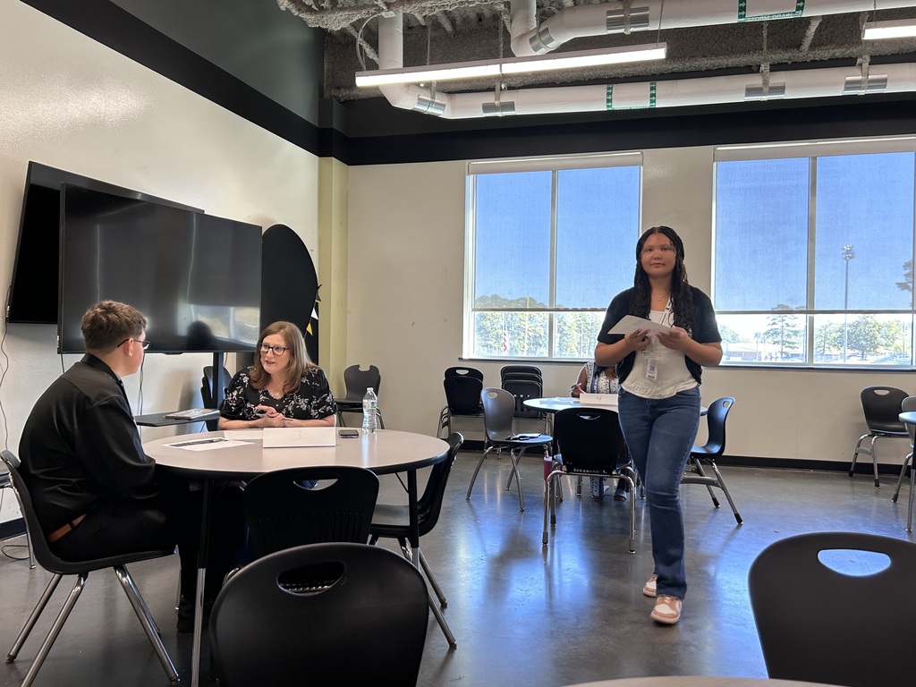 A high school student sitting across a small round table from a adult community interviewer during a mock interview event. The interviewer holds a pen and review documents while speaking with the student. Another student is walking with a paper.