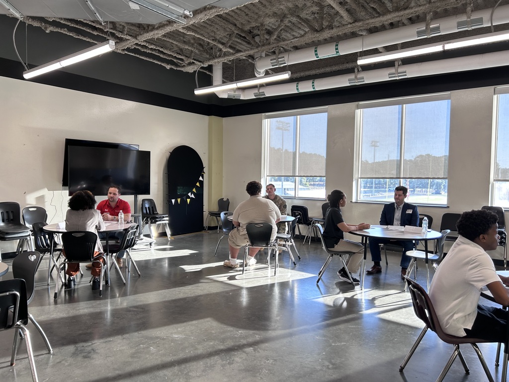 Many high school students sitting across small round tables from a adult community interviewer during a mock interview event. The interviewer holds a pen and review documents while speaking with the student.