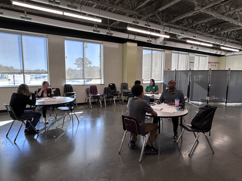Many high school students sitting across small round tables from a adult community interviewer during a mock interview event. The interviewer holds a pen and review documents while speaking with the student.