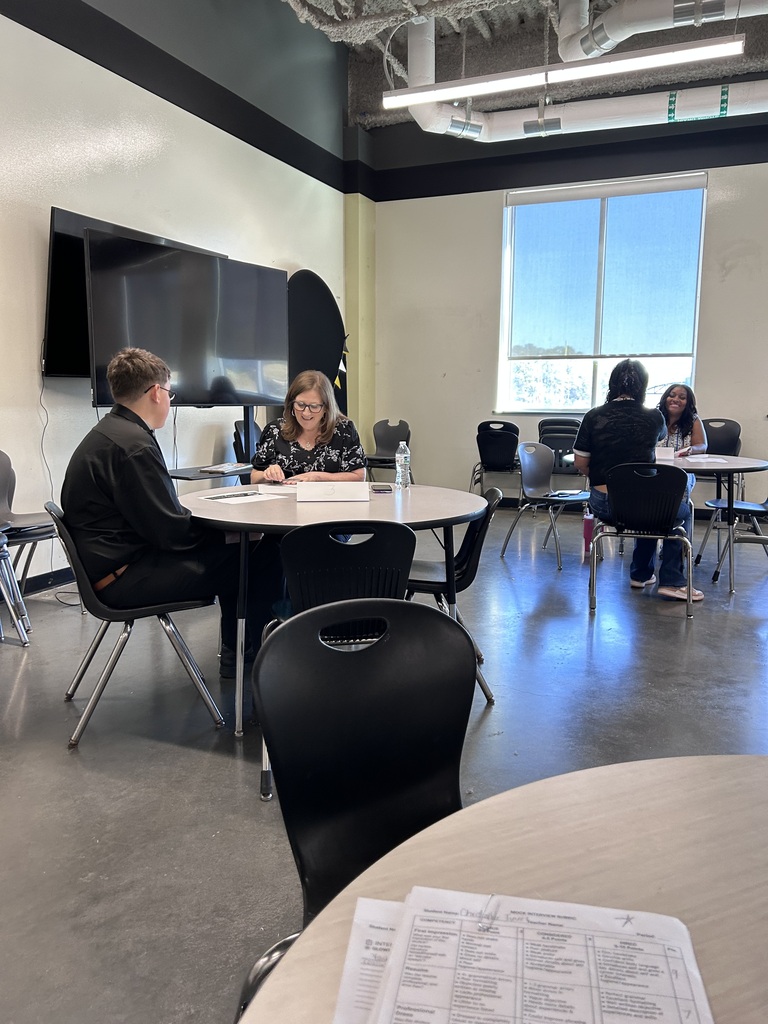 Many high school students sitting across small round tables from a adult community interviewer during a mock interview event. The interviewer holds a pen and review documents while speaking with the student.