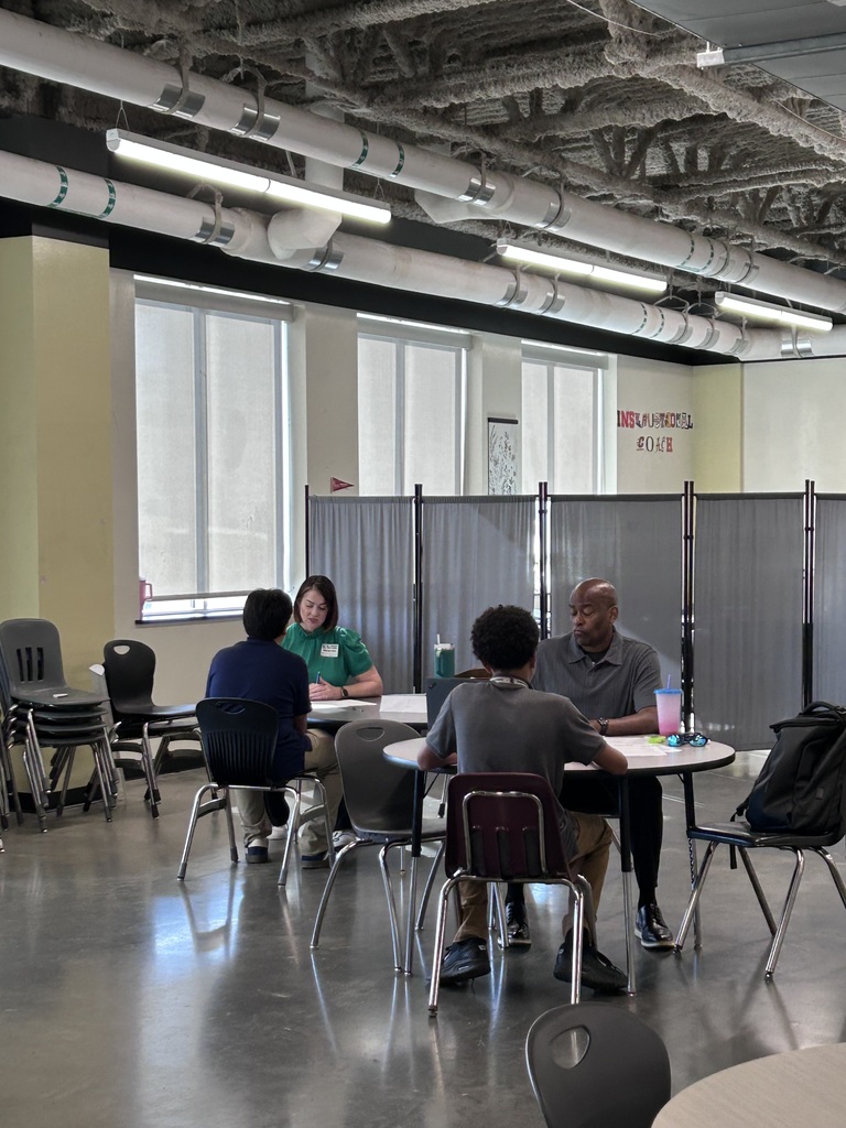 Many high school students sitting across small round tables from a adult community interviewer during a mock interview event. The interviewer holds a pen and review documents while speaking with the student.