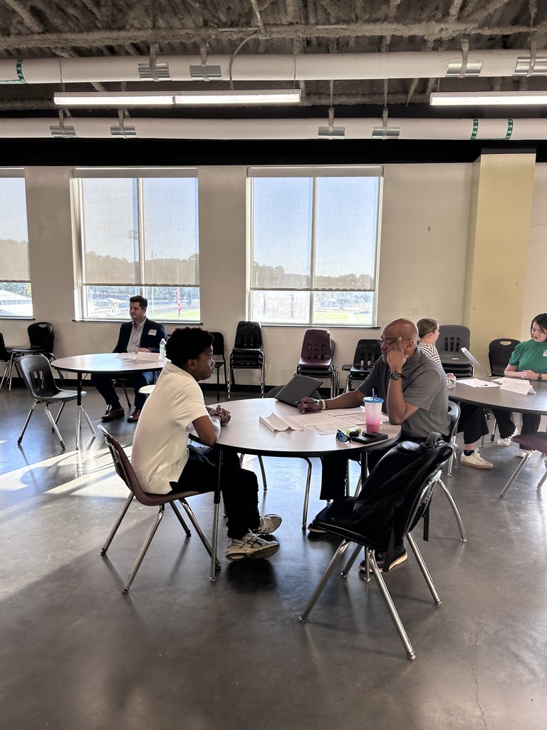 A high school student sitting across a small round table from a adult community interviewer during a mock interview event. The interviewer holds a pen and review documents while speaking with the student.
