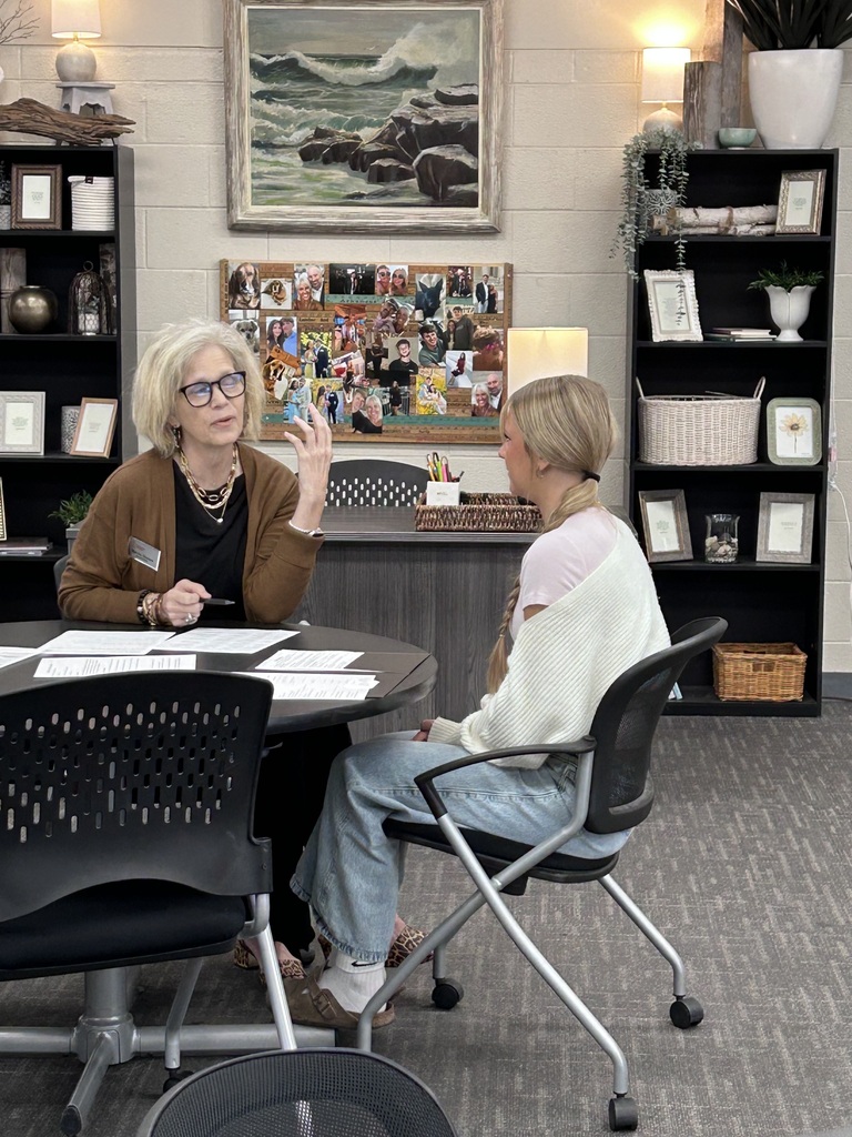 A high school student sitting across a small round table from a adult community interviewer during a mock interview event. The interviewer holds a pen and review documents while speaking with the student.
