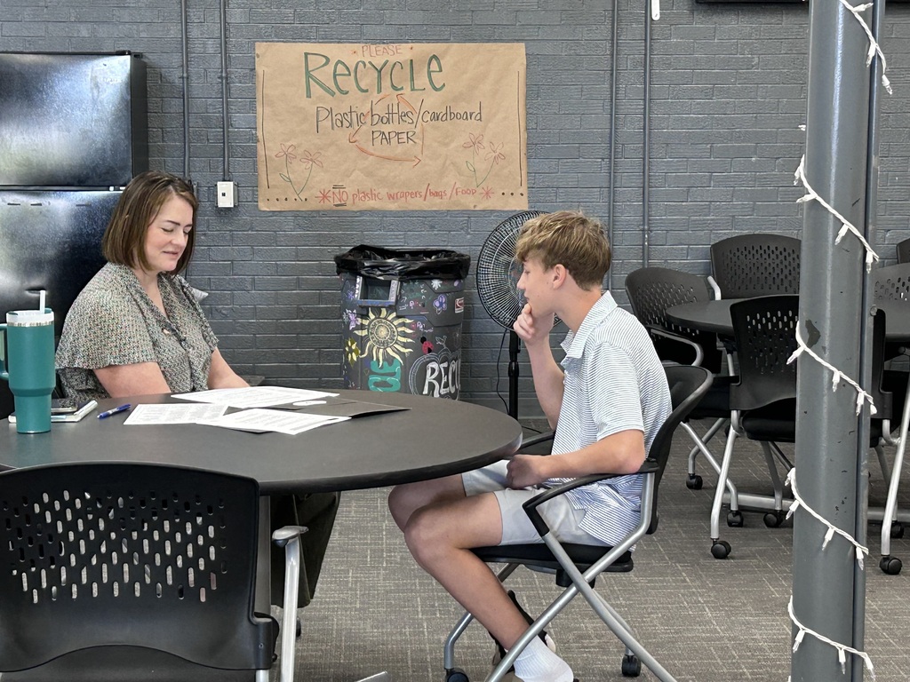 A high school student sitting across a small round table from a adult community interviewer during a mock interview event. The interviewer holds a pen and review documents while speaking with the student.