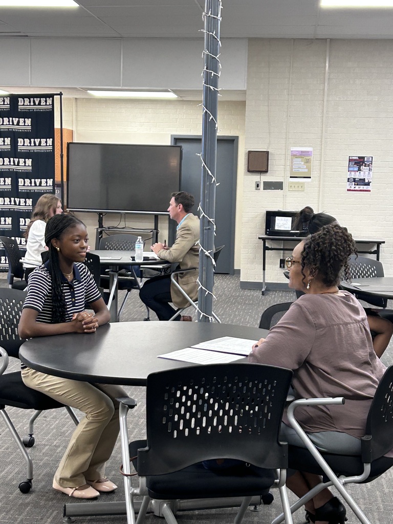 A high school student sitting across a small round table from a adult community interviewer during a mock interview event. The interviewer holds a pen and review documents while speaking with the student.