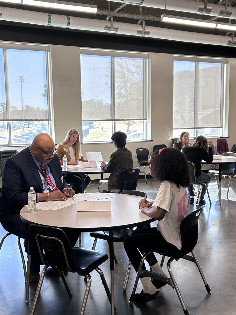 Many high school students sitting across small round tables from a adult community interviewer during a mock interview event. The interviewer holds a pen and review documents while speaking with the student.