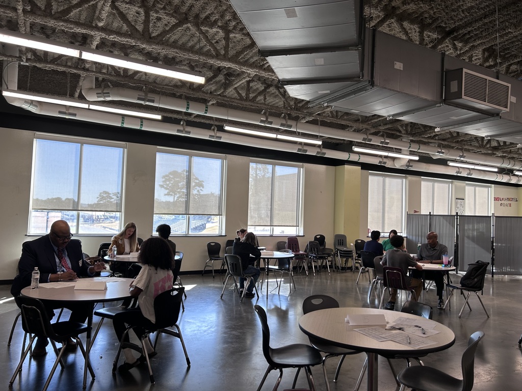 Many high school students sitting across small round tables from a adult community interviewer during a mock interview event. The interviewer holds a pen and review documents while speaking with the student.