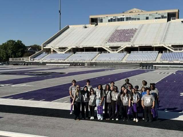 A group of students stands on a university football field during a college visit, holding bags and posing near the sideline as part of a college and career exploration experience.