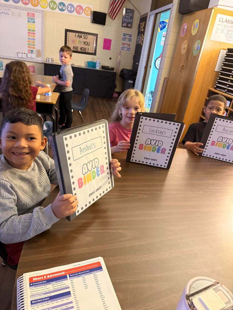 Students seated around a classroom table display their AVID binders during a lesson. Three students hold personalized binders labeled "AVID Binder 2026-2027," highlighting an organizational tool used to support note-taking, goal-setting, and academic success. Classroom learning materials and educational displays are visible in the background.