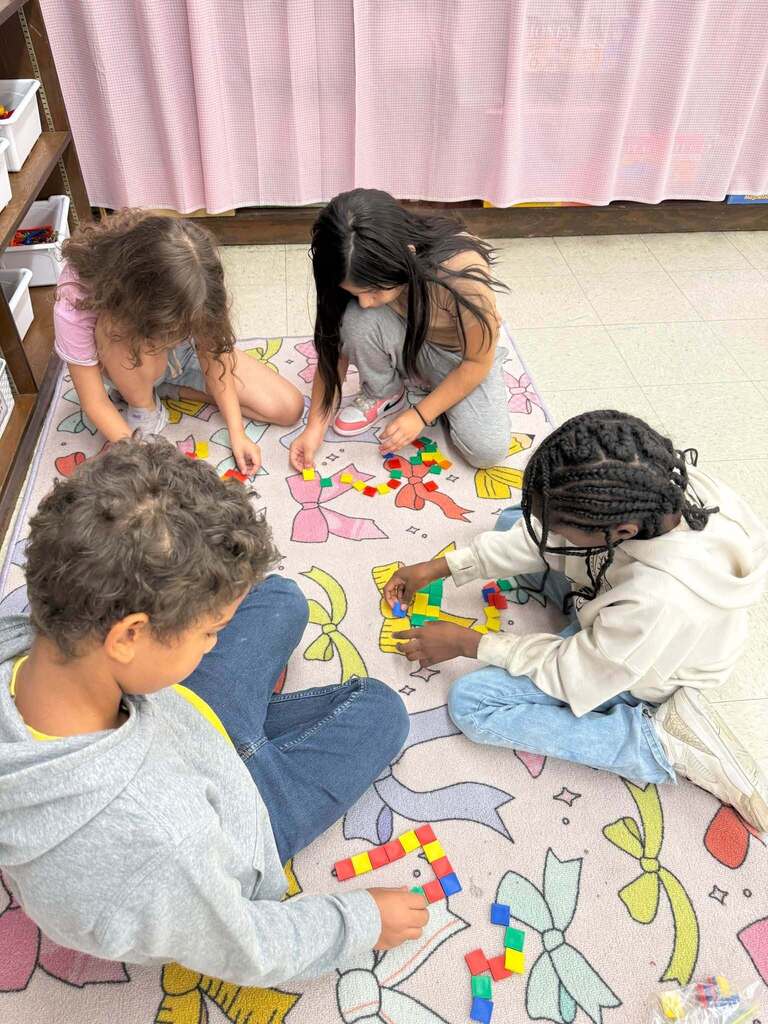 Four students sit on a classroom rug working together with colorful square tiles to create and compare shapes during a collaborative math activity.