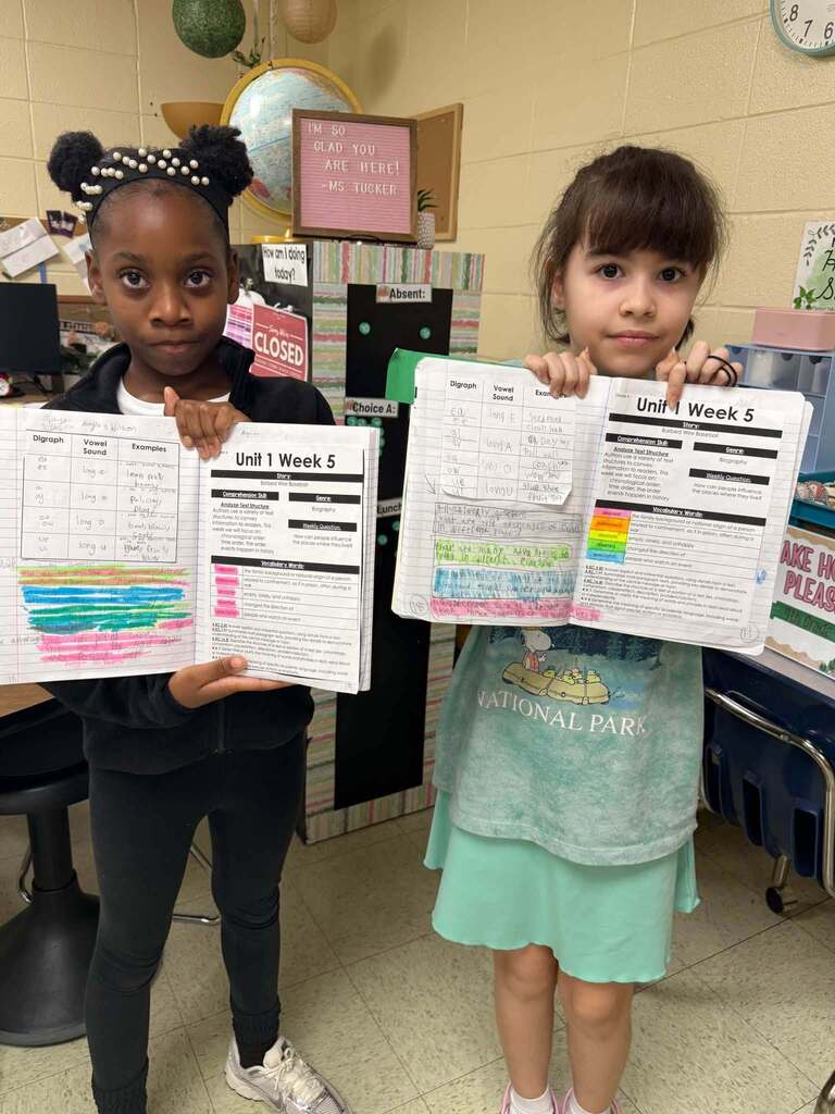 Two students hold open interactive notebooks showing highlighted notes and completed classwork as examples of organized learning and note-taking strategies.