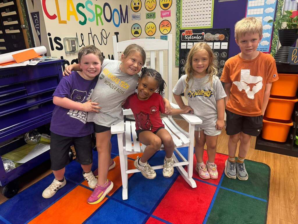 A group of students wearing college-themed shirts gathers around a classroom rocking chair during a college and career readiness activity.