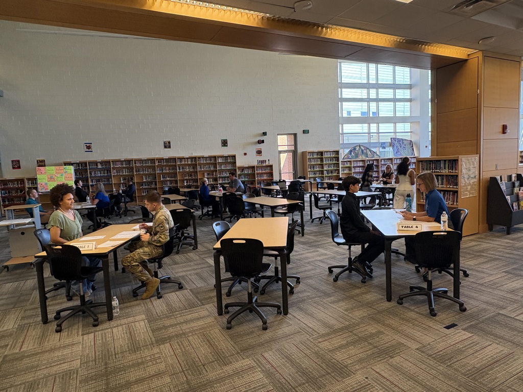 a large library room with many table and chairs. Interviews are being conducted inside between students and adults