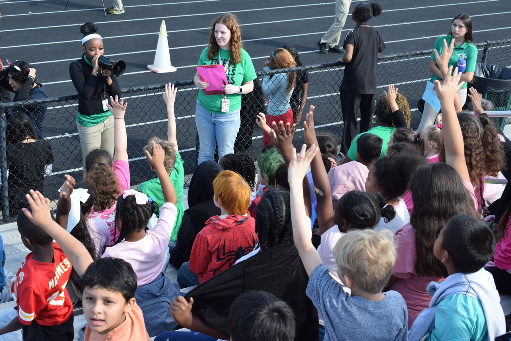 Students waving in bleachers. Older students and teachers are standing in front. One holds a microphone.