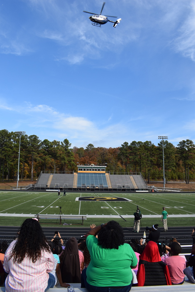 A helicopter taking off in the sky with student and teachers watching from below in stadium bleachers