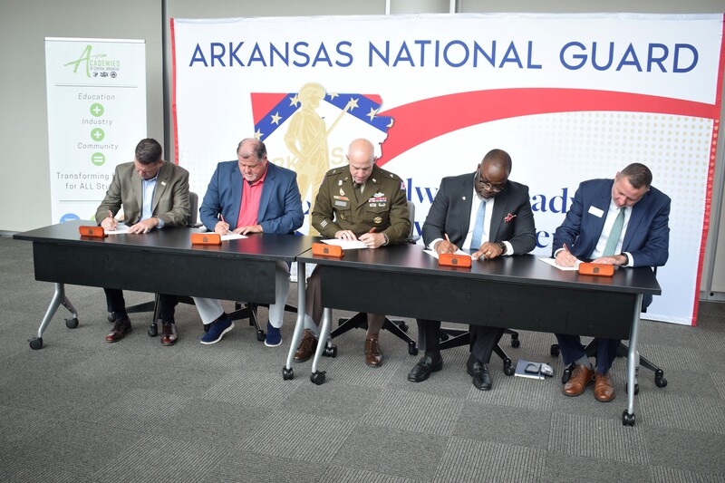 5 men signing at a table with an Arkansas National Guard & Academies of Central Arkansas backdrop behind them