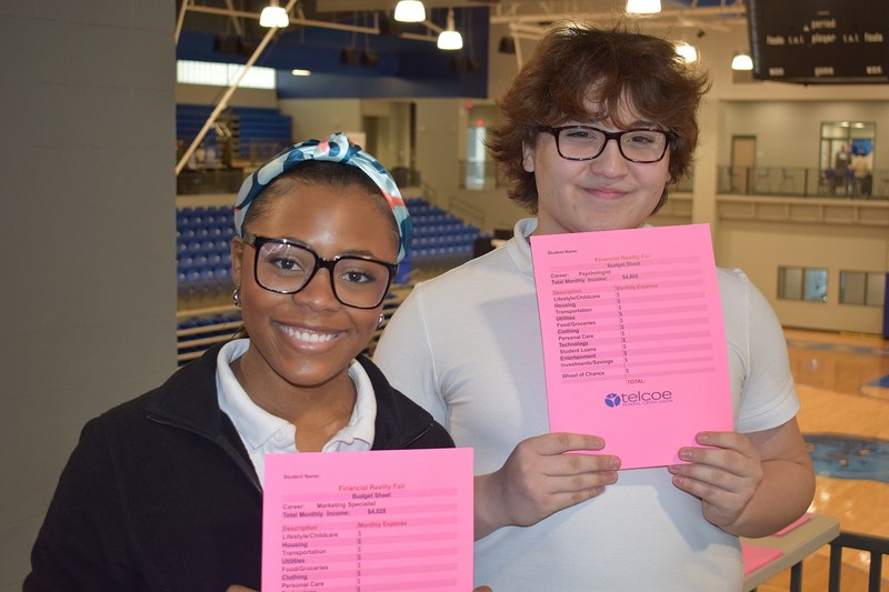 Two students holding Financial Reality Fair papers. The papers have jobs at the top and Telcoe's logo at the bottom.