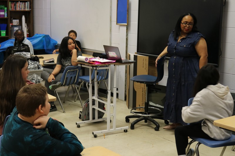 A teacher in front of her students, which are seated in a classroom.