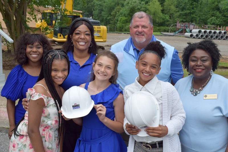 Seven people all wearing blue smile at the camera with construction equipment in the background. Three children are in the front holding hard hats.