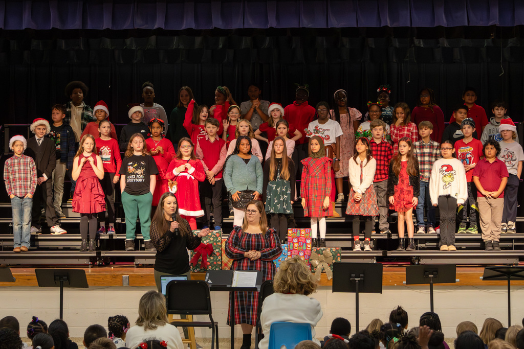 Wide shot of elementary choir standing on risers, two teachers addressing audience