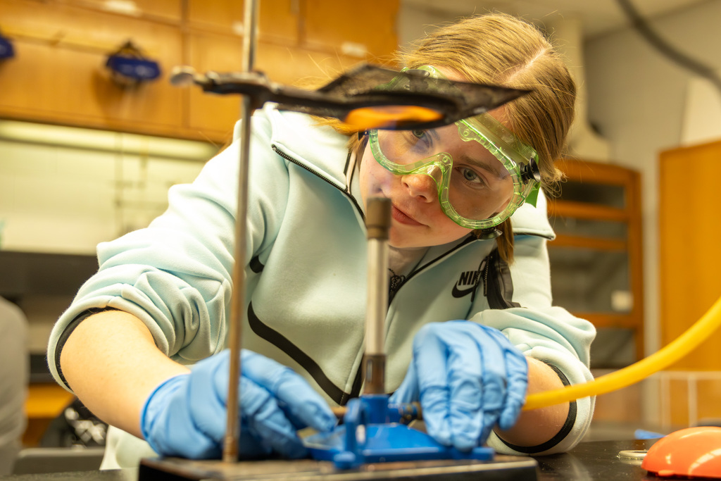 Student wearing goggles and gloves in a science lab lighting a bunsen burner.