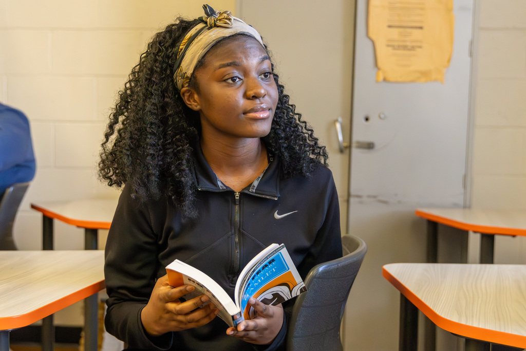 Student sitting in a classroom desk, holding open a copy of Hamlet while listening