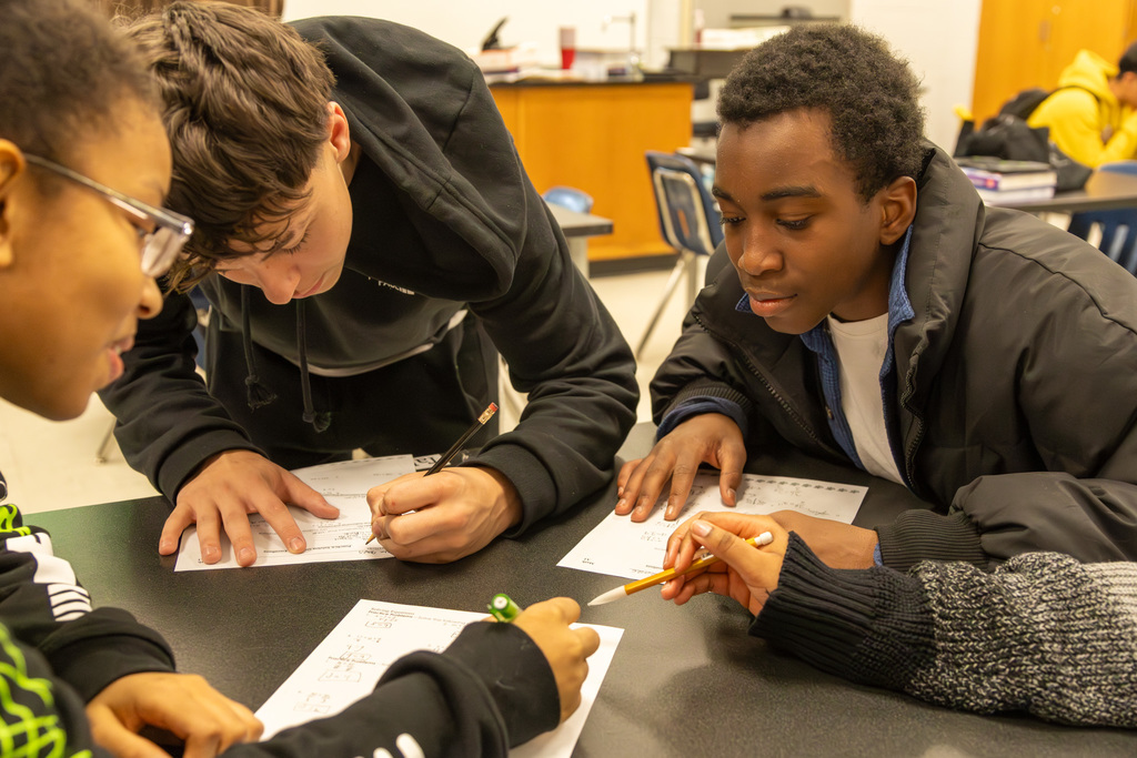 Three students sitting at a lab table looking at worksheets, pointing at each others pages
