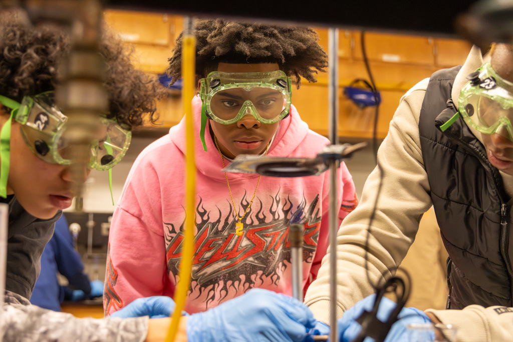 Three students wearing goggles and gloves in a science lab look intently at a bunsen burner flame as they adjust it