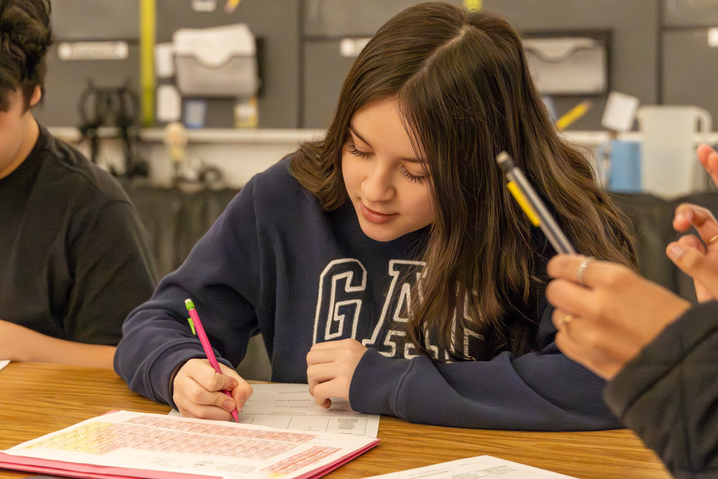 student sitting at a classroom table completing a worksheet with a periodic table