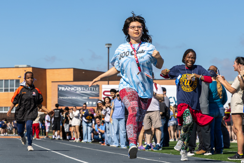 Three students run toward the camera on the track, smiling, as classmates and staff line the sides and cheer them on.