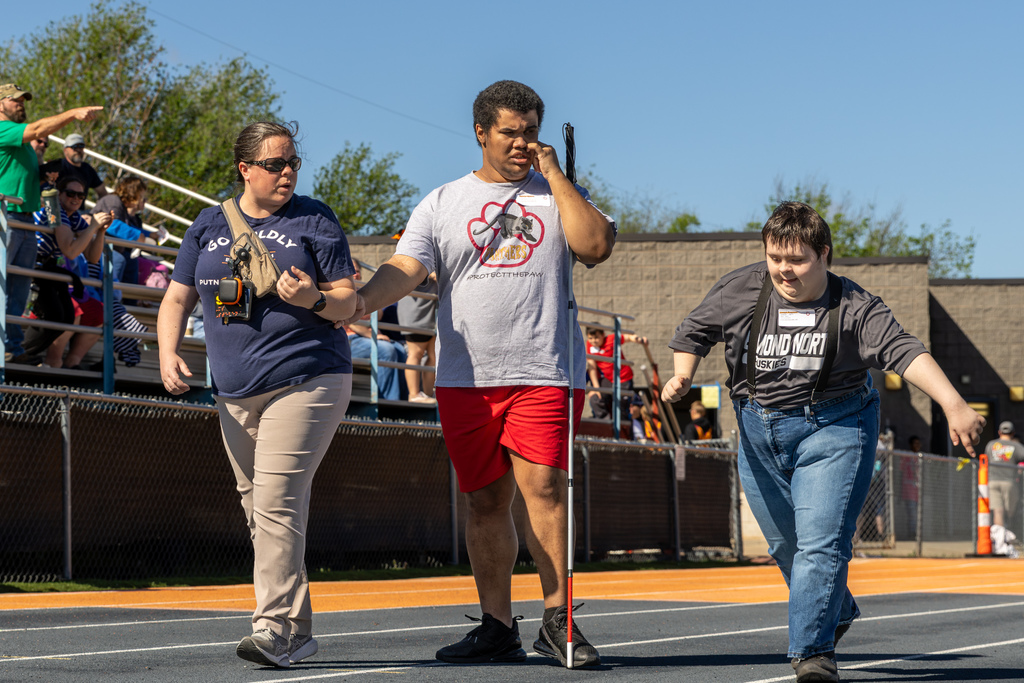 A student walks along the track using a cane with guidance from a staff member, while another student walks alongside them.
