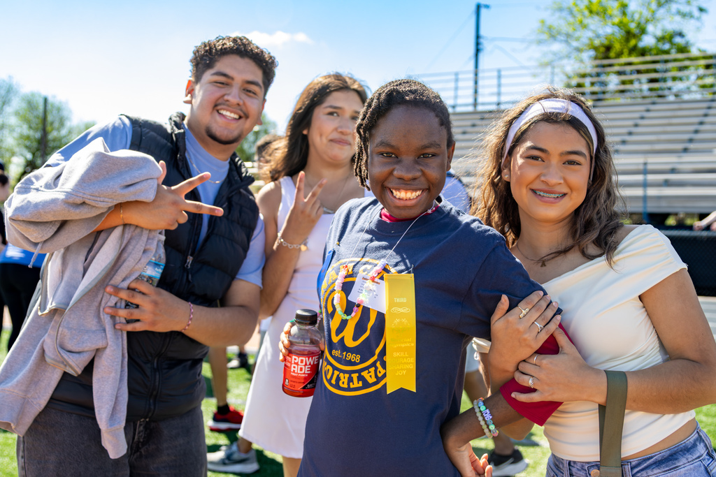 Four students pose together on the field, smiling and flashing peace signs, with one student wearing a ribbon and holding a drink.