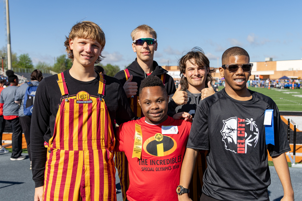 A group of students smiles together on the field, with one student in front wearing a red Special Olympics shirt and a ribbon around his neck.
