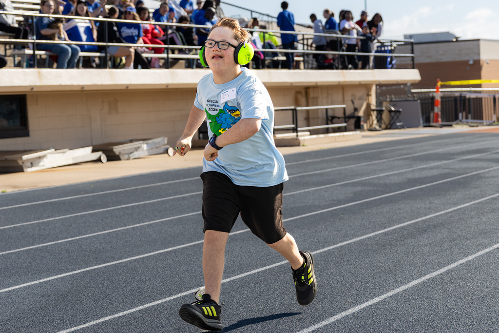A student in a light blue Special Olympics shirt runs down a track lane, wearing bright green headphones, with spectators watching from the stands behind.