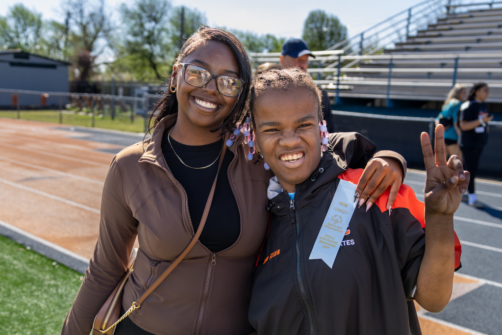 Two students stand side by side on the track smiling at the camera, with one wearing a ribbon and holding up a peace sign.