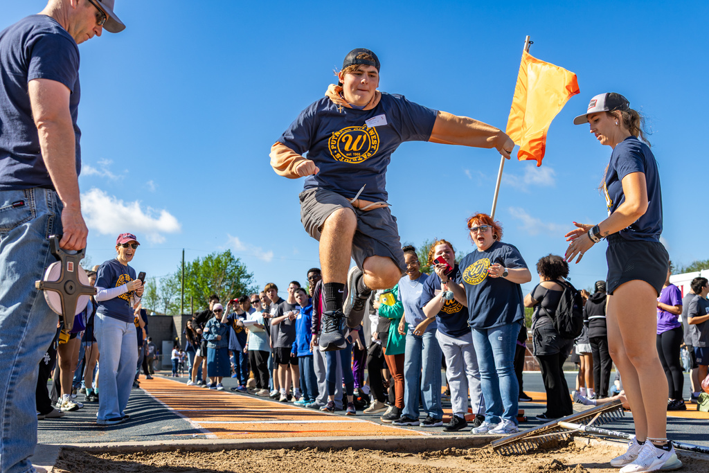 A student leaps into the sand pit during long jump as a crowd lines the runway and cheers from both sides.