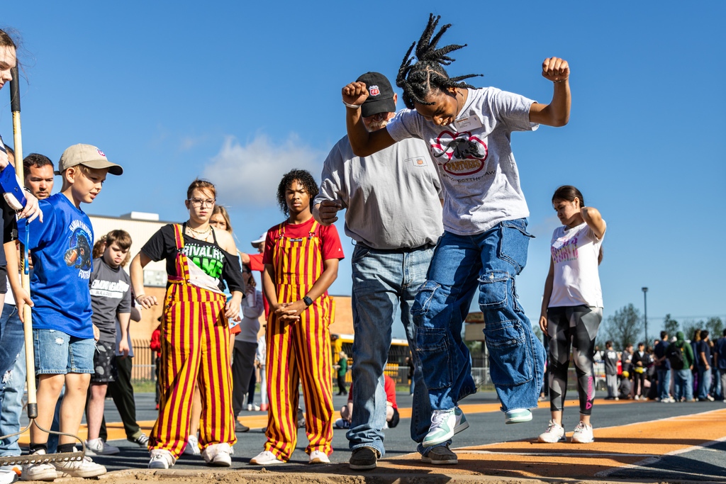 A student lands in the sand during long jump with help from an adult standing beside them, while other students watch from the runway.
