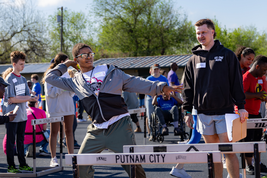 A student winds up and throws a shot put during a field event while a volunteer stands nearby and other students watch in the background.