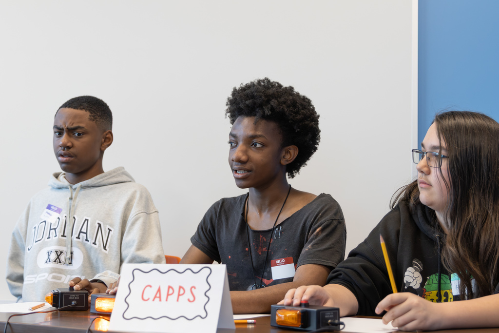 Three students sit side by side at a quiz bowl table labeled “CAPPS,” focused on the question as one student reaches toward the buzzer.