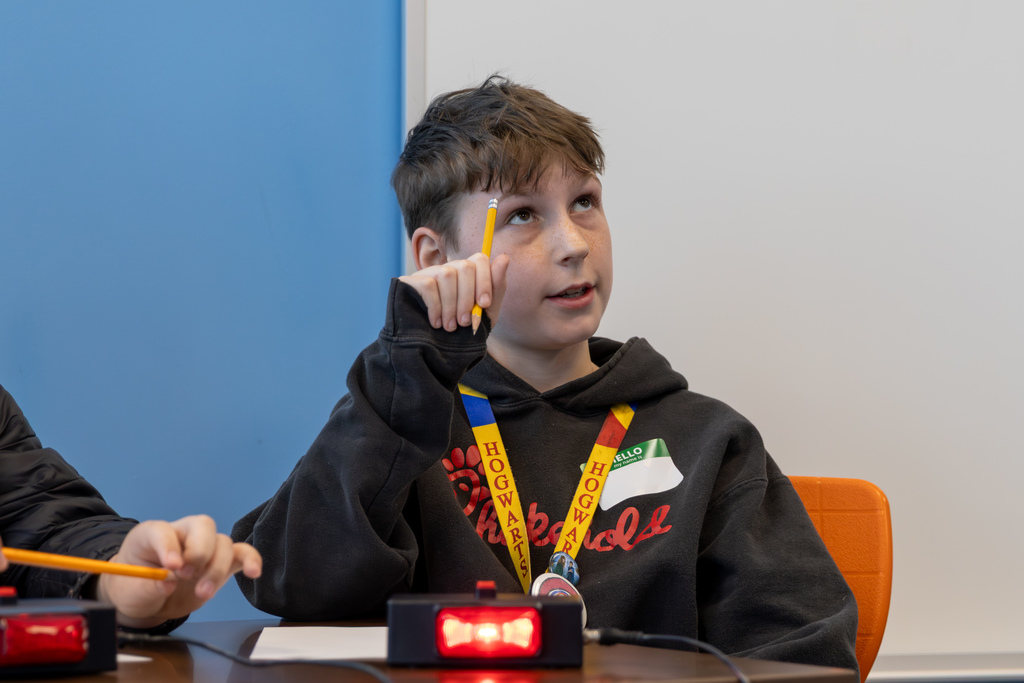A student raises a pencil while thinking through an answer during a quiz bowl round, with the buzzer in front of him.