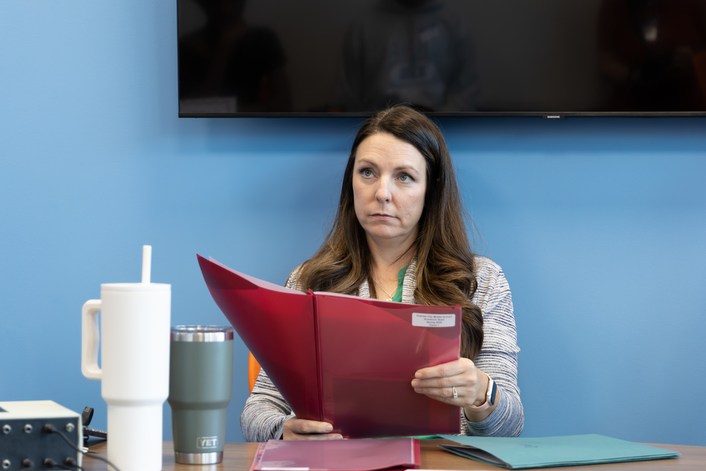 An adult moderator sits at a table holding a red folder, reading questions aloud during a quiz bowl match.