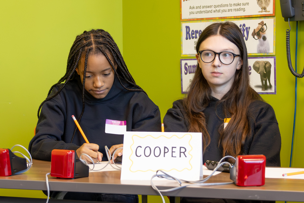 Two students at a table labeled “COOPER” write notes during a quiz bowl match, with buzzers and pencils in front of them.