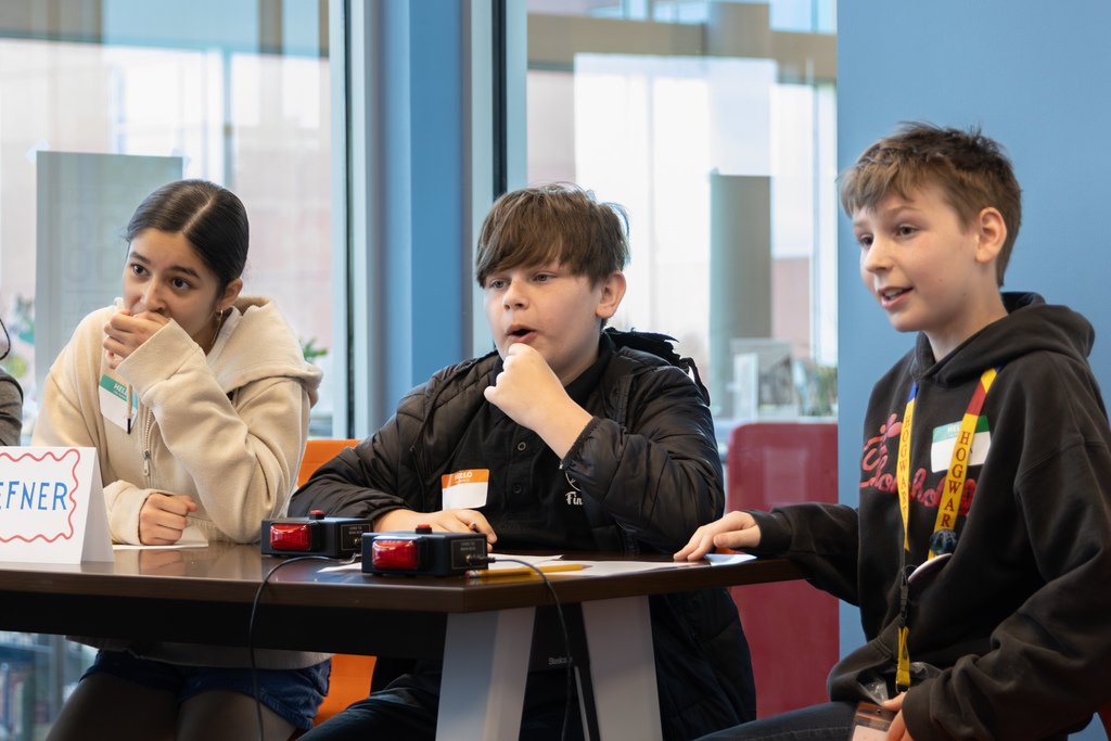 Three middle school students sit at a table during a quiz bowl match, leaning in and watching closely with their hands near the buzzers.