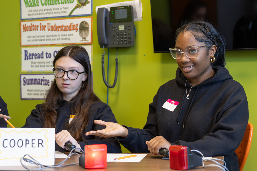 A student gestures while answering a question at a quiz bowl table labeled “COOPER,” as a teammate watches.