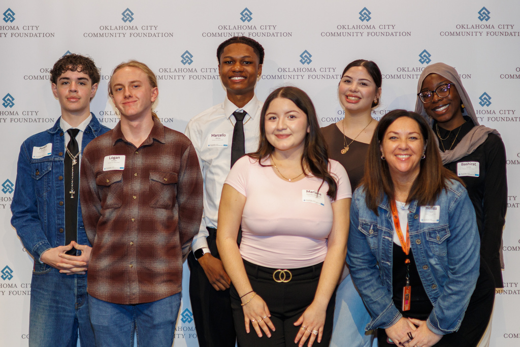 A group of high school students and a staff member stand together smiling in front of an Oklahoma City Community Foundation backdrop during a scholarship banquet.