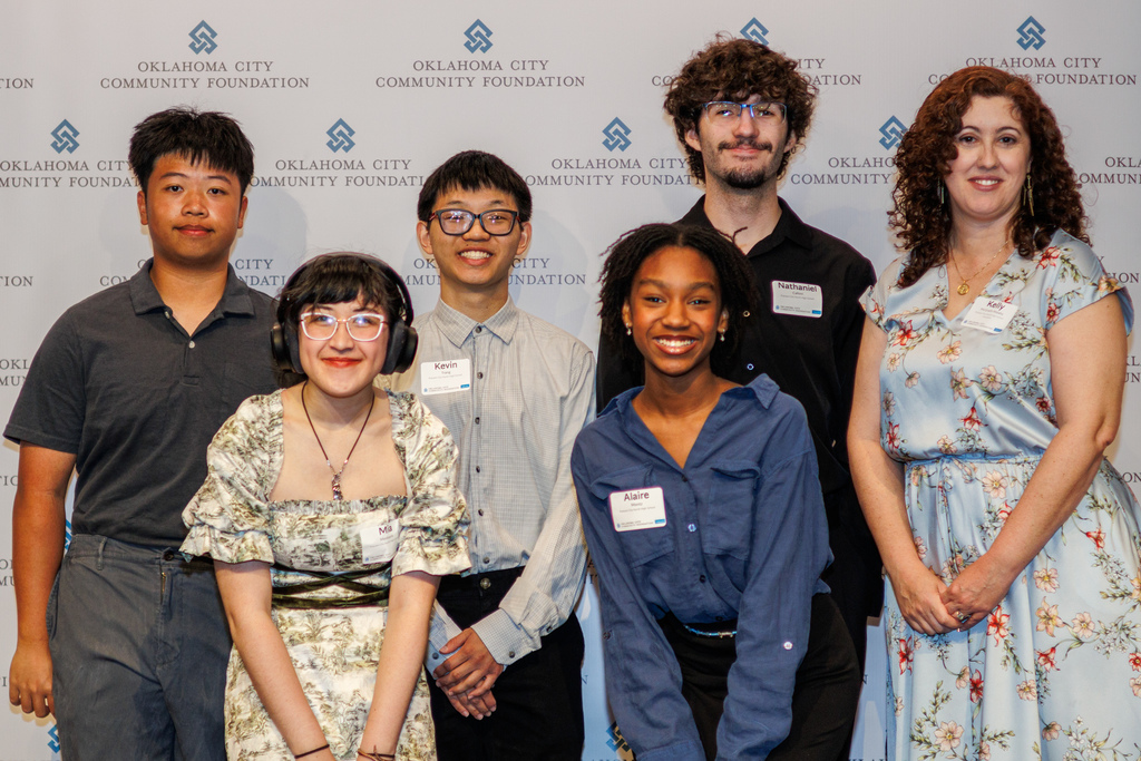 A group of high school students and a staff member stand together smiling in front of an Oklahoma City Community Foundation backdrop during a scholarship banquet.