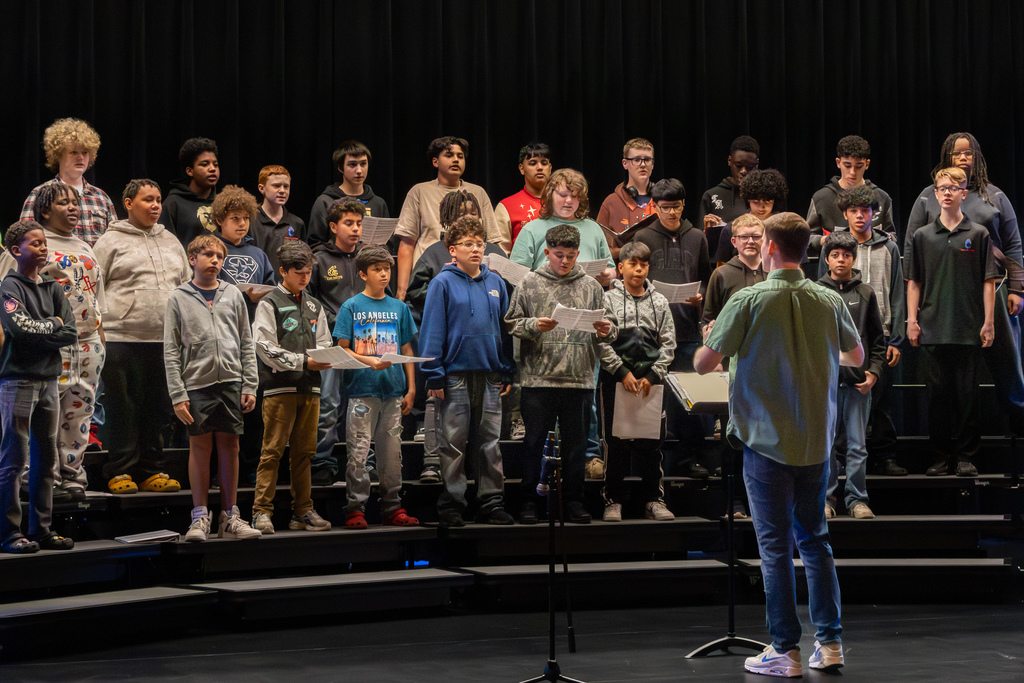 A large group of middle school students stand on risers in a performing arts hall, holding music as a conductor leads them during a tenor-bass choir performance